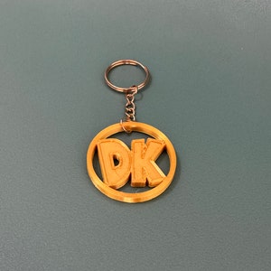 Può includere: Un portachiavi dorato con un ciondolo circolare che presenta le lettere "DK" in stile 3D.