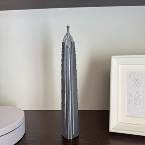 May include: A tall, silver-colored 3D printed model of a skyscraper featuring a tiered, tapering design with a pointed spire at the top. The structure has a vertical, ribbed exterior with multiple levels that decrease in width toward the peak.