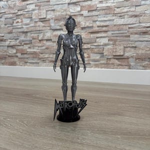 Może przedstawiać: Metalowa, szara figurka żeńskiego robota z filmu "Metropolis". Robot stoi na czarnej podstawie z tytułem filmu. Figurka ma szczegółowy design i jest ustawiona w pozycji pionowej.
