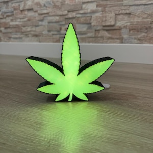 Op de afbeelding: Een groen en zwart nachtlampje in de vorm van een marihuanablad. Het licht is aan en gloeit groen.