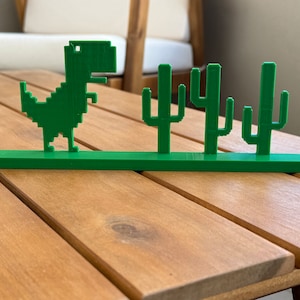 May include: A green 3D printed display featuring a pixelated dinosaur figure standing on a flat base next to three distinct pixelated cactus shapes. The entire object is constructed from a single piece of solid green material with a matte finish.