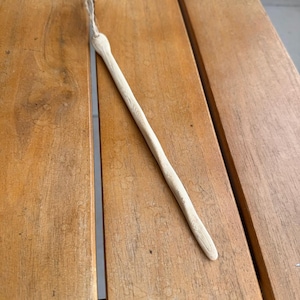 May include: A long, slender, light-colored wooden wand with a smooth, tapered shape and a visible natural grain pattern. A thin, light-tan twine loop is attached to the rounded top end of the wooden stick, allowing for easy hanging or display.