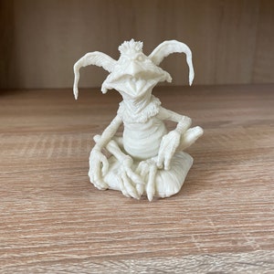 May include: A white 3D printed figurine of a creature with large ears, a wide smile, and long fingers. The creature is sitting on a small cushion.