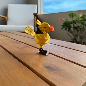 May include: A yellow origami figure of a Chocobo, a bird-like creature from the Final Fantasy video game series, standing on a wooden table.
