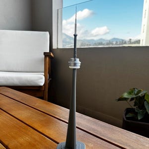 Peut inclure: Une miniature grise de la Tour CN, un célèbre monument de Toronto, au Canada. Le modèle est fait d'un matériau gris foncé et repose sur une base grise. La tour est sur une table en bois avec vue sur un ciel bleu et des montagnes en arrière-plan.
