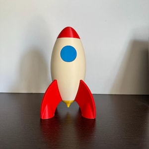 May include: A toy rocket ship featuring a cream-coloured body, a bright red nose cone, and three red stabilising fins at the base. The rocket includes a circular blue window on the front and a small yellow pointed thruster at the bottom centre.