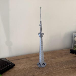 May include: A silver-coloured 3D printed model of the Tokyo Skytree tower. The structure features a tall, slender, tapering design with a textured lattice exterior, a circular observation deck section, and a thin antenna spire at the very top.
