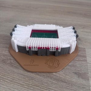 May include: A 3D-printed model of a stadium, featuring a green field, white seating, and a brown base. The base includes the text "MiniWorld3D" and a team logo. The stadium model is a detailed replica.