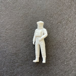 May include: A small, white, three-dimensional figurine of an astronaut in a full-body space suit. The figure features a detailed helmet, a chest-mounted control panel, and ribbed textures along the arms and legs, standing in a neutral pose.