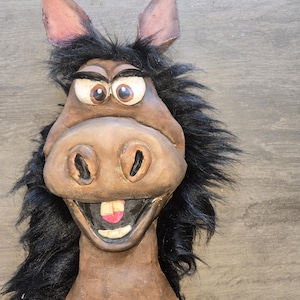 May include: A whimsical donkey mask with a brown face, large expressive eyes, and a wide, toothy grin. The mask features black faux fur around the face and ears, and is likely made of a sturdy material.