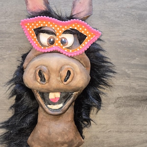 May include: A whimsical donkey mask with large, expressive eyes and a playful grin. The mask features a bright orange and pink polka-dot eye mask and black faux fur mane. The donkey has large ears and a detailed, textured face.