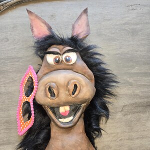 May include: A whimsical donkey mask with large, expressive eyes, a wide grin, and black fur. A pink and orange polka-dot mask rests on the side. The mask is made of a brown material.