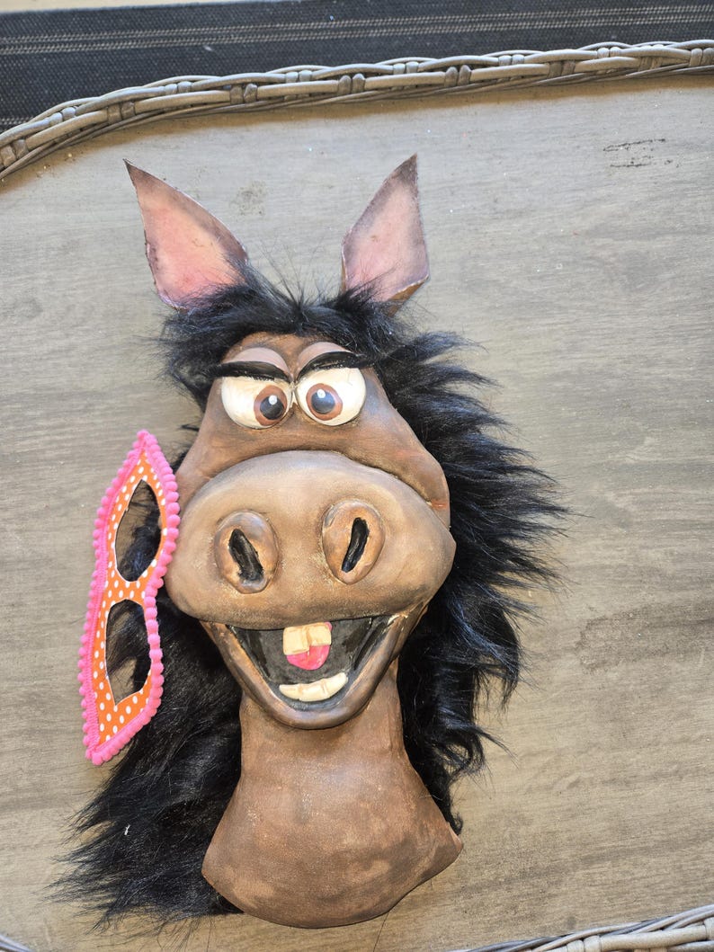 May include: A whimsical donkey mask with large, expressive eyes, a wide grin, and black fur. A pink and orange polka-dot mask rests on the side. The mask is made of a brown material.