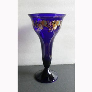 Könnte beinhalten: Eine tief kobaltblaue Glasvase mit ausgestelltem Rand und einem Standfuß. Der obere Teil der Vase ist mit einem aufwendigen Zierband in Gold, Weiß und Hellblau verziert, das aus wirbelnden Mustern und kreisförmigen Motiven besteht.