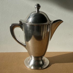 Könnte beinhalten: Eine silberne Kanne mit einem Scharnierdeckel und einem runden Knopf oben. Die Kanne hat einen hohen, schlanken Körper und einen ausgestellten Boden.