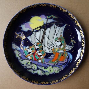 Peut inclure: Une assiette décorative avec un fond bleu foncé et un bord doré. L'assiette présente une illustration colorée d'un bateau avec des personnages, une pleine lune et des nuages. Le bateau a une voile blanche et navigue sur une mer de nuages.