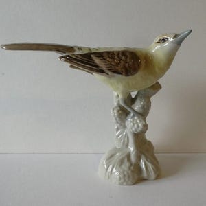 May include: A ceramic bird ornament, perched on a decorative branch. The bird features a pale yellow body, brown wings, and a long tail. The branch and base are white, contrasting with the bird's colours. The ornament is a decorative item.