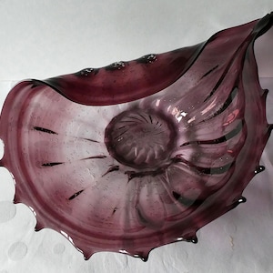 May include: A large, purple glass bowl with a wavy, organic shape and a textured surface. The bowl has a wide, open mouth and a deep, rounded base.