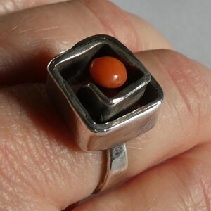 May include: A silver ring featuring a square-shaped setting with a smooth, polished finish. Centred within the square frame is a small, rounded, vibrant orange stone that sits slightly recessed, creating a layered and dimensional appearance.