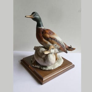 Könnte beinhalten: Keramik-Entenfigur. Ein männlicher Erpel mit grünem Kopf und braunem Körper sitzt über einer kleineren Ente. Die Enten stehen auf einem weißen Sockel, der auf einem rechteckigen Holzsockel befestigt ist. Dekorativer Artikel.