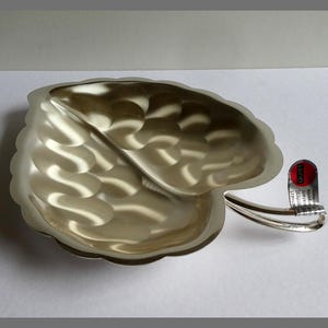May include: A silver-coloured leaf-shaped dish with a textured surface. The dish has a stem-like handle and a small tag attached. The leaf design features detailed veining and a scalloped edge.