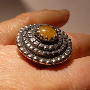 Könnte beinhalten: Ein silberfarbener Ring mit einem großen, ovalen, bernsteinfarbenen Stein in der Mitte. Der Ring hat ein geschichtetes Design mit einem strukturierten, geriffelten Muster um den Stein. Der Ring befindet sich an einem Finger.