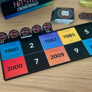 May include: A board game with a black game board featuring numbered squares and colorful cards with song titles and years. The game includes circular tokens and a box labeled "HITSTER". The game board is on a wooden surface.