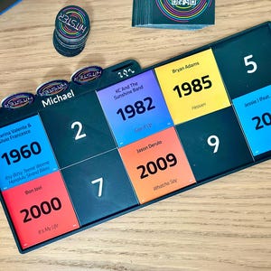 May include: A board game with colorful tiles featuring song titles and artist names. The game includes circular tokens and a stack of cards. The tiles display years like 1960, 1982, 1985, 2000, 2009, and 2011. The game is set on a wooden surface.