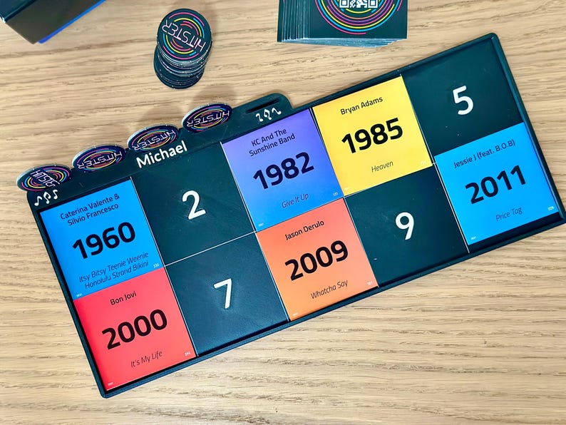 May include: A board game with colorful tiles featuring song titles and artist names. The game includes circular tokens and a stack of cards. The tiles display years like 1960, 1982, 1985, 2000, 2009, and 2011. The game is set on a wooden surface.