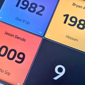 May include: Close-up of a colorful grid featuring song titles and years. The squares display the years 1982, 1985, and 2009, along with song titles like "Give It Up," "Heaven," and "Whatcha Say." The background is a dark blue.