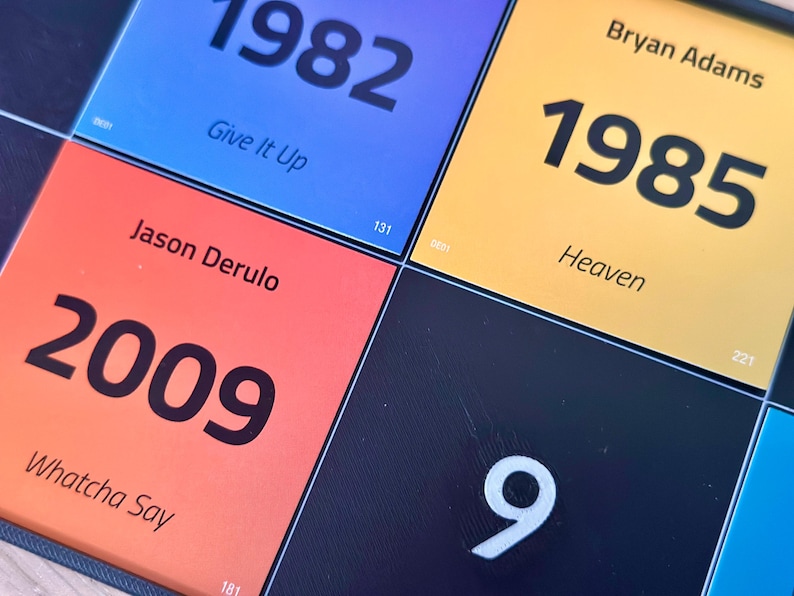 May include: Close-up of a colorful grid featuring song titles and years. The squares display the years 1982, 1985, and 2009, along with song titles like "Give It Up," "Heaven," and "Whatcha Say." The background is a dark blue.