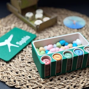 Könnte beinhalten: Ein grüner Brettspiel-Organizer mit bunten Spielsteinen und Spielmarken. Der Organizer hat Schlitze für Spielmarken mit verschiedenen Designs. Eine grüne Karte mit weißem Text und einer weißen Grafik ist ebenfalls vorhanden.