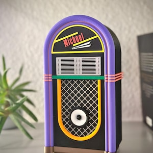 Op de afbeelding: Een decoratief jukeboxmodel met een zwarte behuizing, paarse boogvormige randen, oranje accenten en een zilverkleurig luidsprekerrooster. Het bovenste gedeelte toont de naam Michael in rode letters op een zwarte achtergrond met gele lijnen.