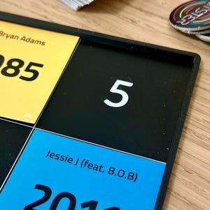 May include: A black game board with yellow, blue, and black squares. The yellow square has the text "Bryan Adams 1985 Heaven". The blue square has the text "Jessie J (feat. B.O.B) 2011". A white number "5" is on a black square. Colorful circular game pieces are in the background.