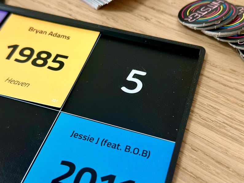 May include: A black game board with yellow, blue, and black squares. The yellow square has the text "Bryan Adams 1985 Heaven". The blue square has the text "Jessie J (feat. B.O.B) 2011". A white number "5" is on a black square. Colorful circular game pieces are in the background.