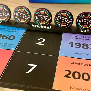 May include: A black board game with colorful squares featuring song titles and years. Five circular HITSTER buttons with a neon design are on top. The name "Michael" is written on the board. The game includes songs from 1960, 1982, and 2009.