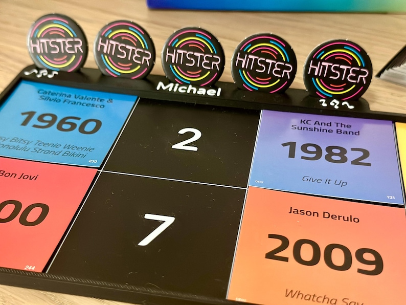 May include: A black board game with colorful squares featuring song titles and years. Five circular HITSTER buttons with a neon design are on top. The name "Michael" is written on the board. The game includes songs from 1960, 1982, and 2009.
