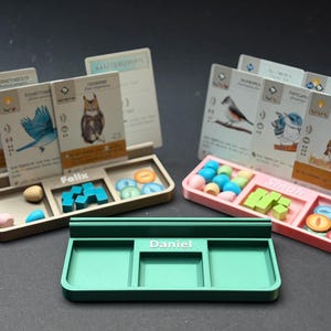 May include: Three game component trays in beige, pink, and green, each with three compartments. The trays hold game pieces like wooden cubes, round tokens, and small spheres. Game cards with bird illustrations are displayed behind the trays.