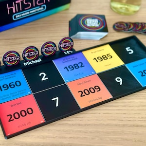 May include: A board game with a black game board featuring colorful squares with years and song titles. The game includes circular tokens with the word "HITSTER" and a box with the game's title. The game is set on a wooden surface.