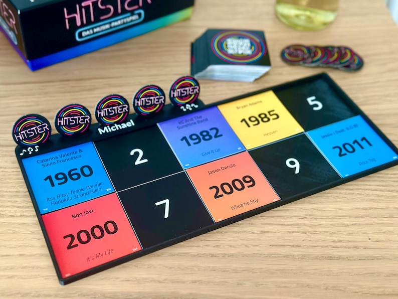 May include: A board game with a black game board featuring colorful squares with years and song titles. The game includes circular tokens with the word "HITSTER" and a box with the game's title. The game is set on a wooden surface.