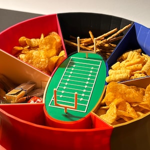 Könnte beinhalten: Ein mehrfarbiger, stadionförmiger Serviertablett mit sechs Fächern. Das Tablett zeigt ein grünes Fußballfeld mit weißen Yardlinien und einem Tor im Zentrum. Die Fächer sind mit verschiedenen Snacks gefüllt, darunter Chips, Brezeln und Cracker.