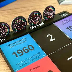 May include: A black game board with colorful cards and circular tokens. The tokens have the word "HITSTER" in neon colors. The board features the year 1960, the name "Michael", and the number "2".