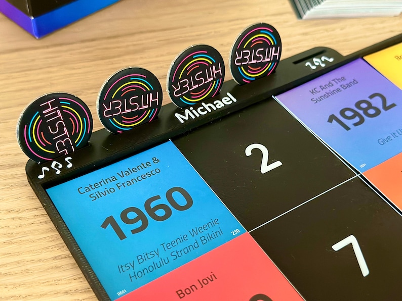 May include: A black game board with colorful cards and circular tokens. The tokens have the word "HITSTER" in neon colors. The board features the year 1960, the name "Michael", and the number "2".