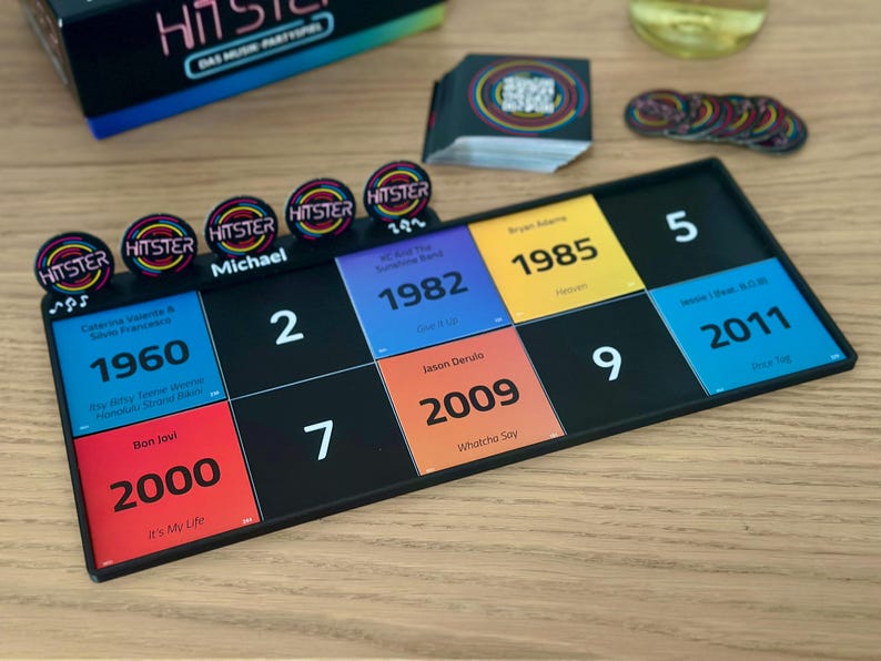 May include: A board game with a black game board featuring numbered squares and colorful cards with song titles and years. The game includes circular tokens and a box labeled "HITSTER". The game board is on a wooden surface.