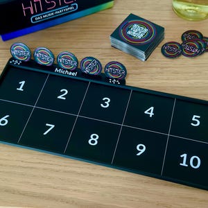 May include: A black board game set with the word "HITSTER" on the game pieces and box. The game board is divided into ten numbered squares. There are game pieces, cards, and a box, all with colorful accents. The game is set on a wooden surface.