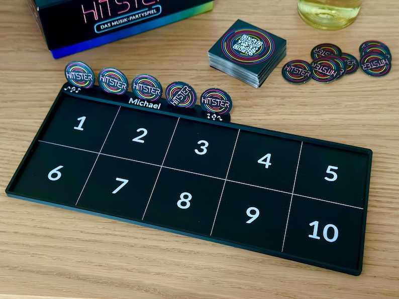 May include: A black board game set with the word "HITSTER" on the game pieces and box. The game board is divided into ten numbered squares. There are game pieces, cards, and a box, all with colorful accents. The game is set on a wooden surface.