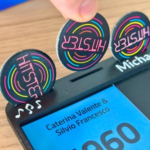 May include: A black game piece with the word "HITSTER" in pink neon, surrounded by concentric circles of yellow, blue, and green. The game piece is being placed in a black holder with the name "Michael" printed on it.