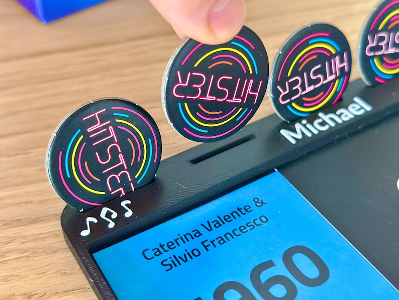 May include: A black game piece with the word "HITSTER" in pink neon, surrounded by concentric circles of yellow, blue, and green. The game piece is being placed in a black holder with the name "Michael" printed on it.