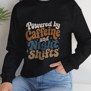 以下が含まれることがあります： 「Powered by Caffeine and Night Shifts」の文字が白、茶色、青でプリントされた黒いスウェットシャツ。長袖でクルーネックのデザイン。デザインはシャツの前面中央に配置されています。