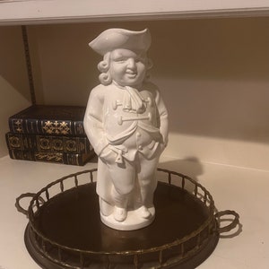 May include: A white porcelain figurine of a man wearing a tricorn hat and a jacket with buttons. The figurine is standing on a round brass tray with a bamboo-style rim.